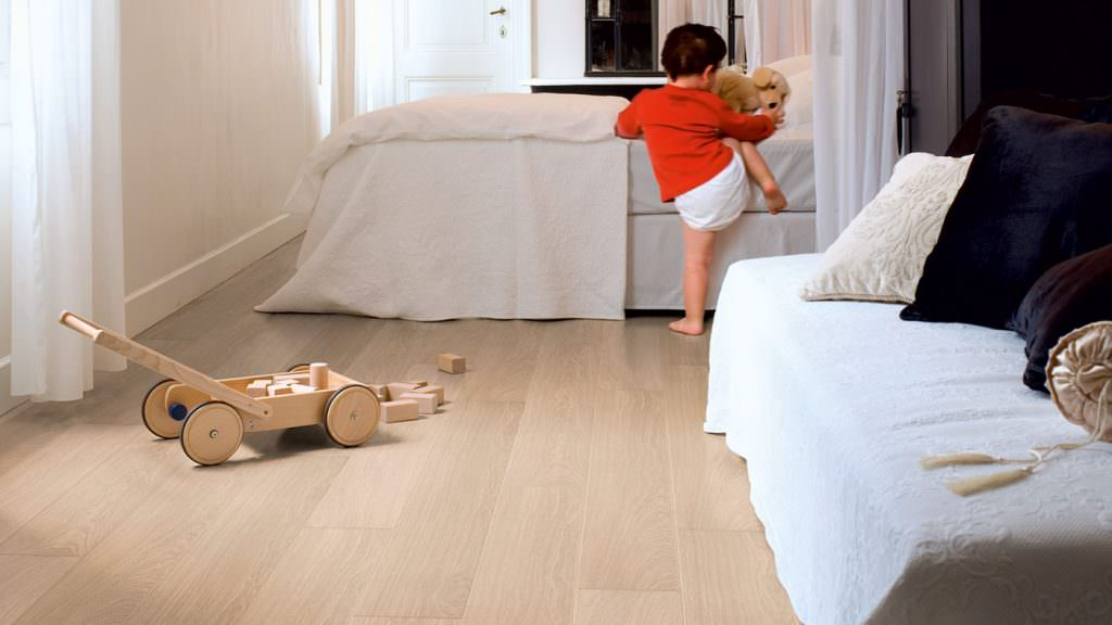 Criança subindo na cama para brincar com o ursinho, apoiando o pé esquerdo no piso laminado.