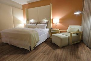 Quarto de Casal com piso vinílico amadeirado instalado junto de uma cama e e um sofá cama e cortina com paredes alaranjadas.